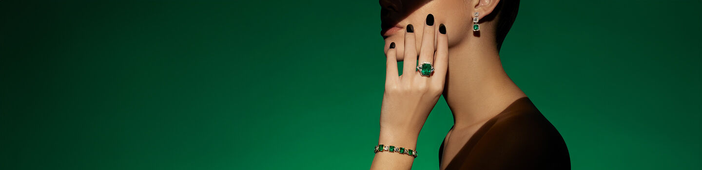 Amodel wearing an emerald ring, bracelet and earrings from the Maison Birks high jewellery collection on a green backround.