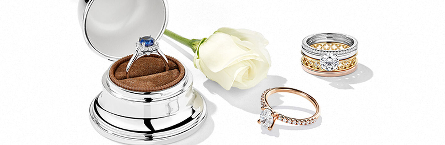 Sapphire ring in a bell box beside a tri-colour ring and a white rose