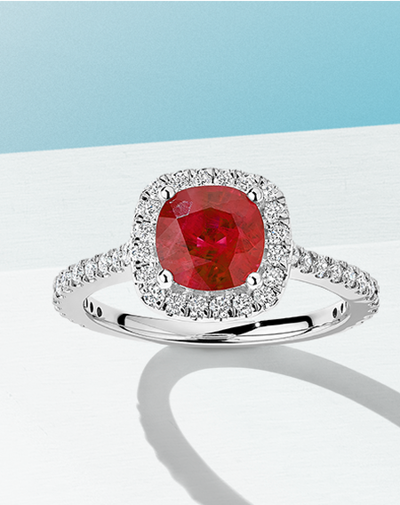 Salon ruby ring with diamond halo and pavé band on a white and light blue background.