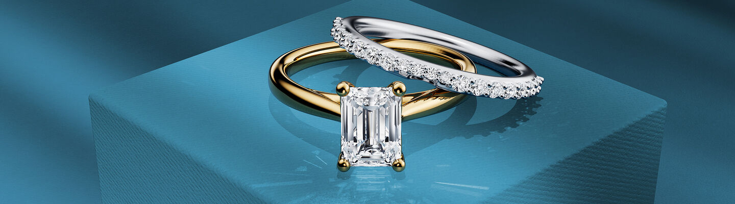 Birks 1879 solitaire emerald ring and diamond wedding band on a blue background.