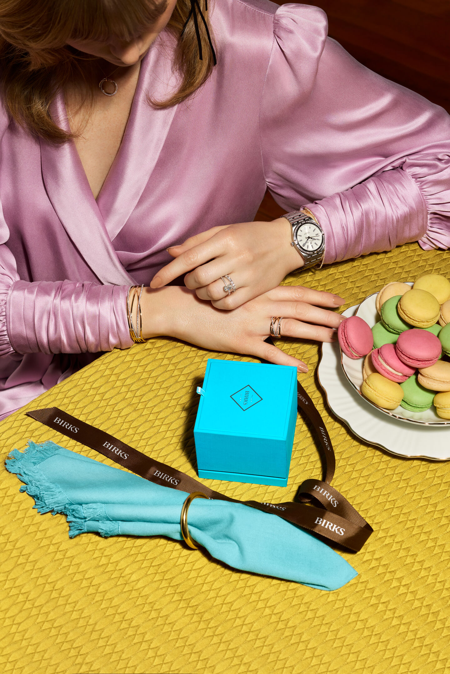 A woman sitting at the dining table Birks blue box and macaroon decorations wearing Birks Ros&eacute;e du Matin  jewellery collection and Breitling Choronomat timepiece.