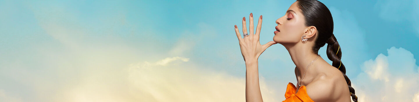 A woman wearing Messika jewellery in an orange dress with a blue background with clouds.