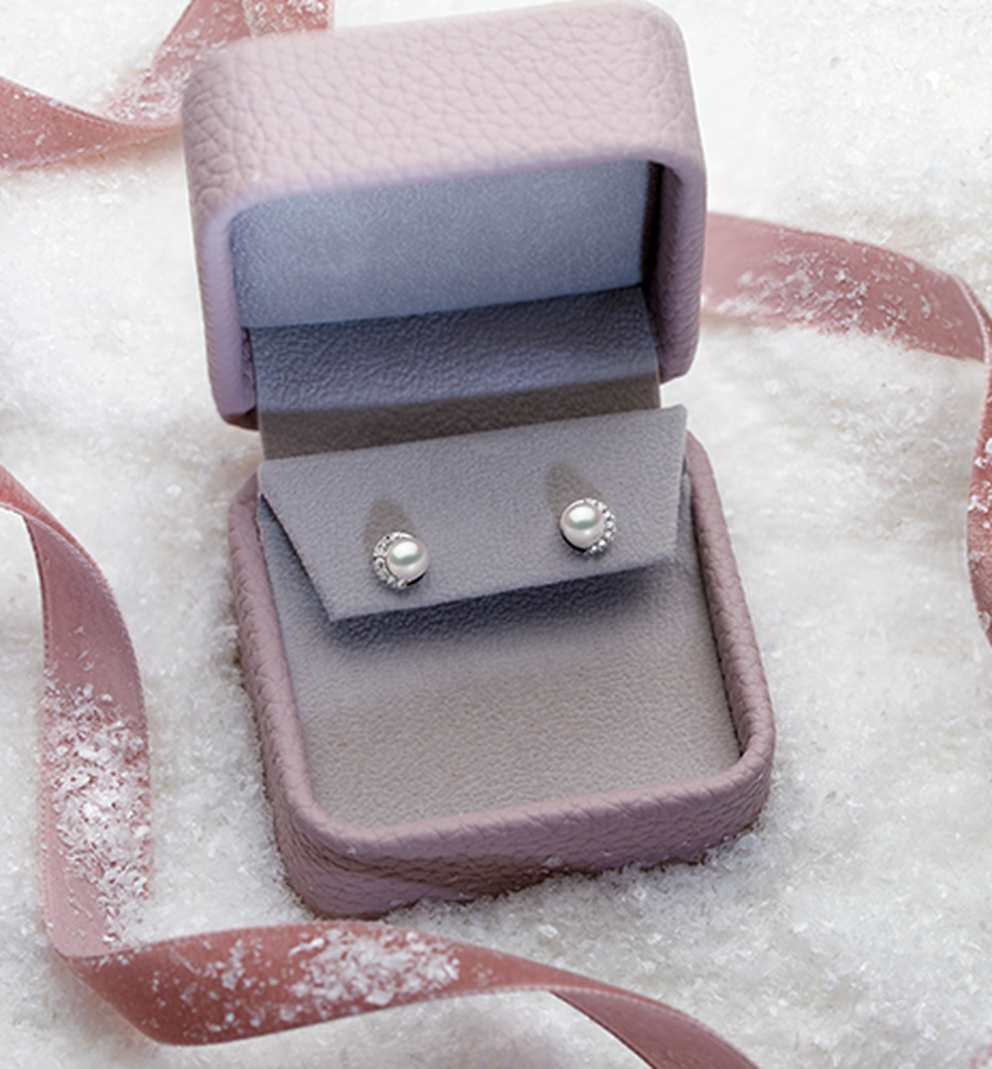 A box containing Yoko London pearl and diamond earrings rest on a pile of snow with a pink ribbon.