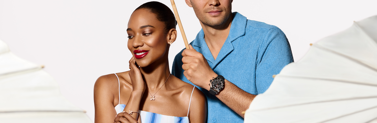 A couple under the umbrella; woman wearing Pasquale Bruni petit garden, petit joli and petit luce collection in earring, necklace and ring, man wearing a Breitling chronomat watch on a beige background.