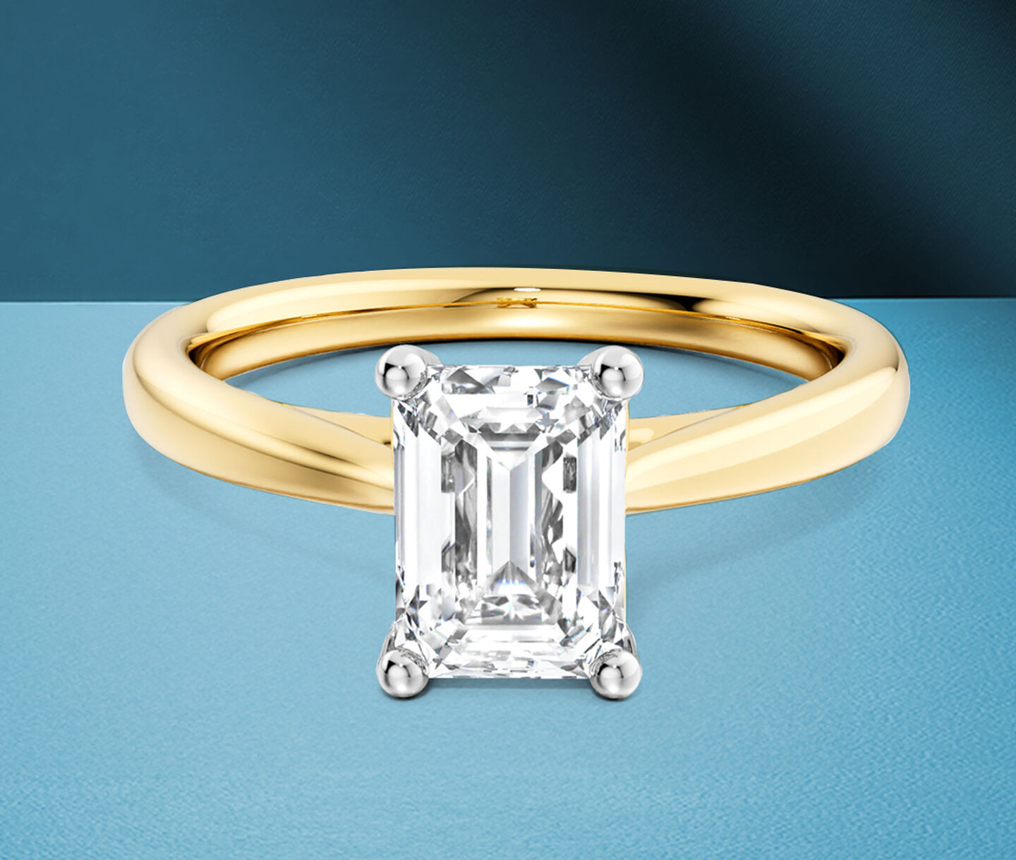 Birks 1879 diamond emerald-cut engagement ring in yellow gold on a blue background.