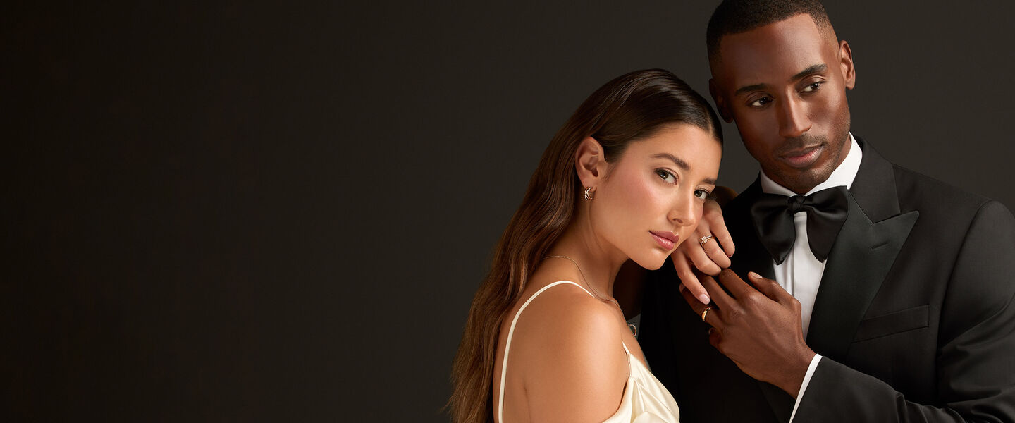 An elegant couple showcasing a Birks engagement ring, gold wedding band, and fine jewellery including a pair of diamond earrings and a necklace, perfect for weddings and special occasions.