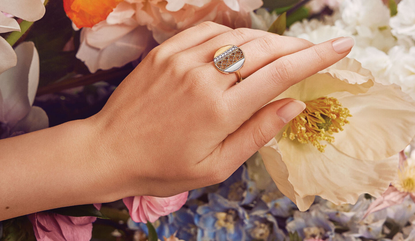 A hand modeling the Birks Dare to Dream circle ring with flowers in the background.