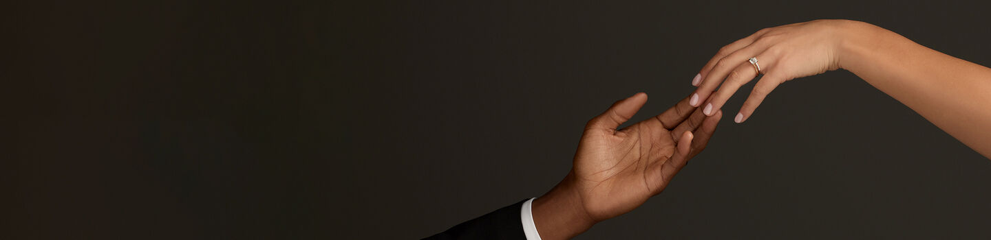 Close-up of two hands reaching toward each other against a dark background. One hand in a black suit sleeve is extended palm up, while the other, adorned with a Birks engagement ring, gently touches it.